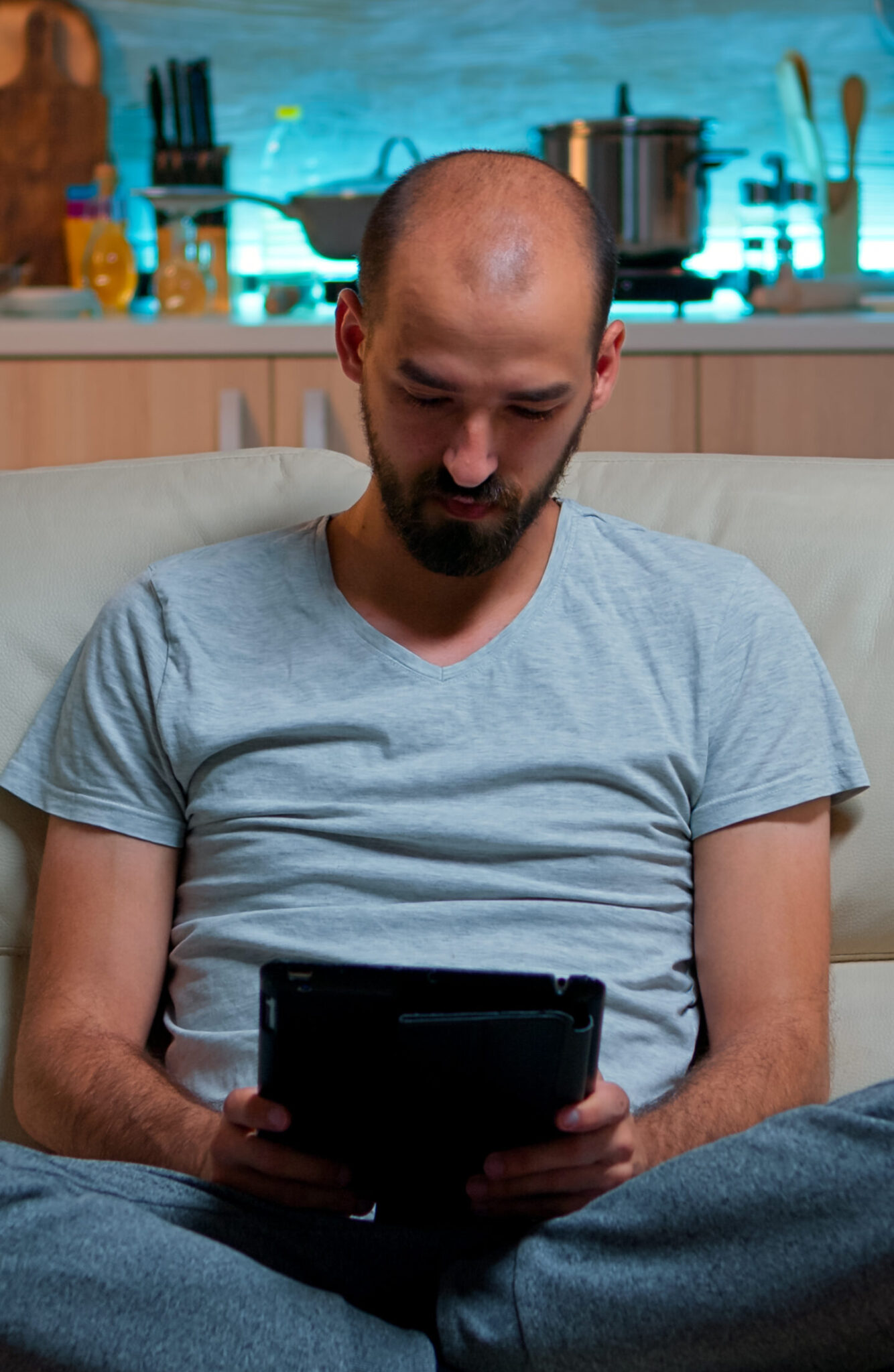 Tired man sitting alone on couch while browsing on internet using tablet computer with modern technology wireless. Caucasian male in pajamas relaxing in front of televion late at night in kitchen
